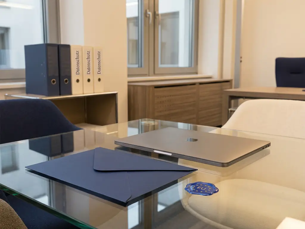 Versiegelter Briefumschlag und Laptop auf Glasschreibtisch mit EU-Datenschutzsiegel und Datenschutz-Ordnern im Hintergrund.