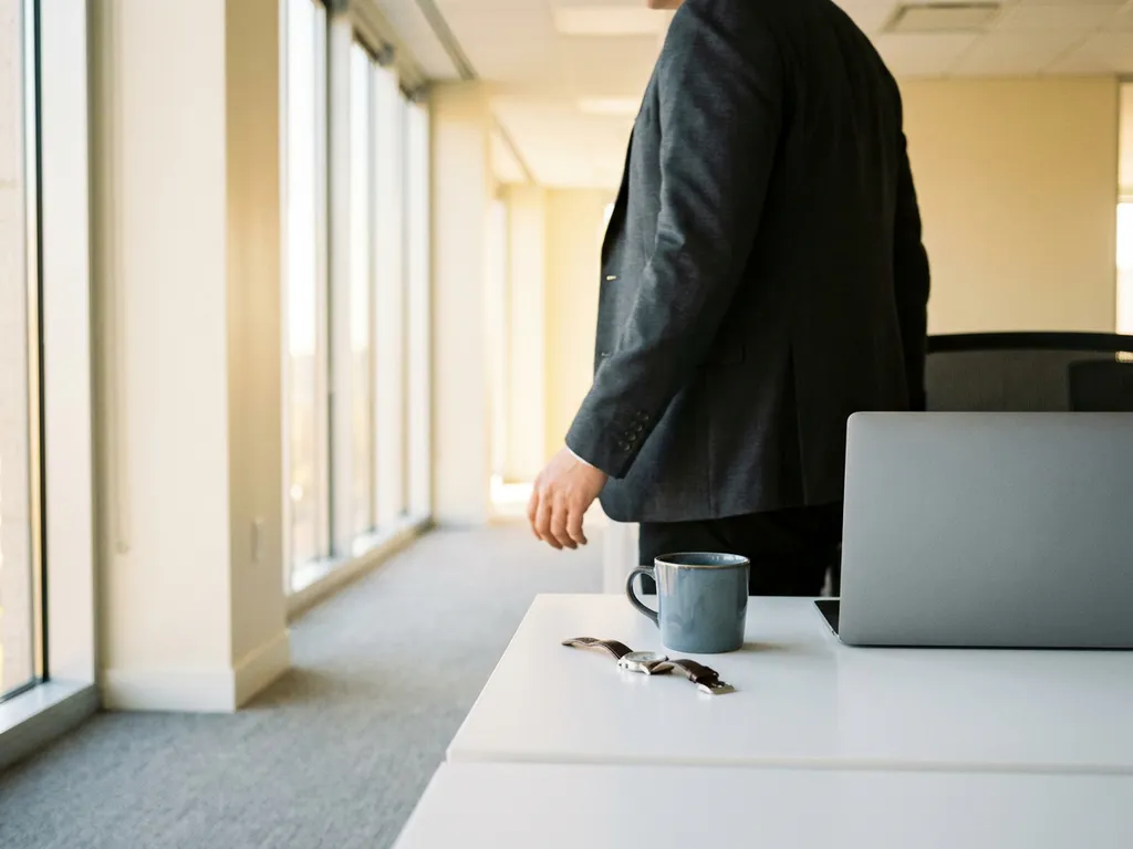 Fachkraft verlässt modernen HR-Software-Arbeitsplatz; Uhr und Kaffeetasse auf weißem Schreibtisch, sonnendurchfluteter Büroflur im Hintergrund.